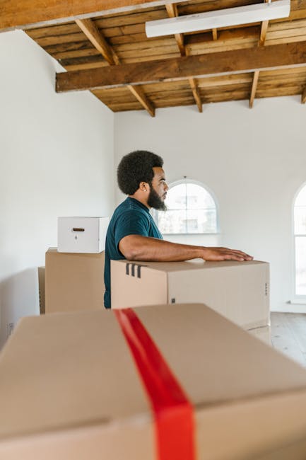 Two women are preparing for a home relocation inside a bright living room with large arched windows allowing natural light to fill the space. One woman, with curly black hair and a white T-shirt, stan